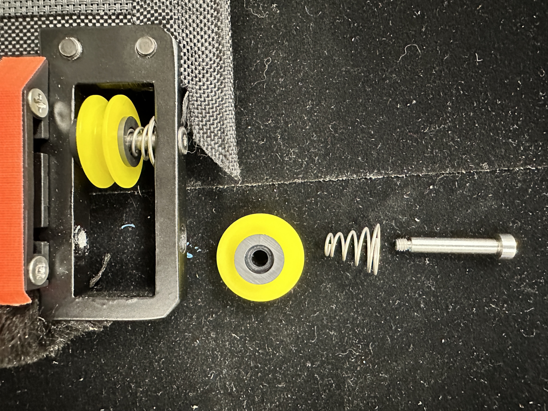 Disassembled KAOS roller bracket showing yellow wheel, coil spring, and bolt laid out separately beside assembled bracket with dual yellow wheels and spring loaded axle